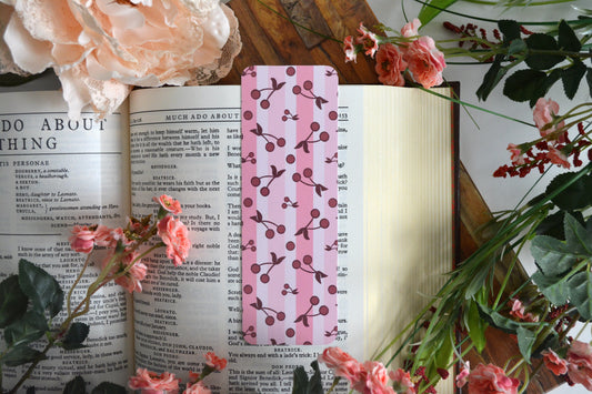 Bookmark with illustrations of cherries on a striped background of varying shades of pink.